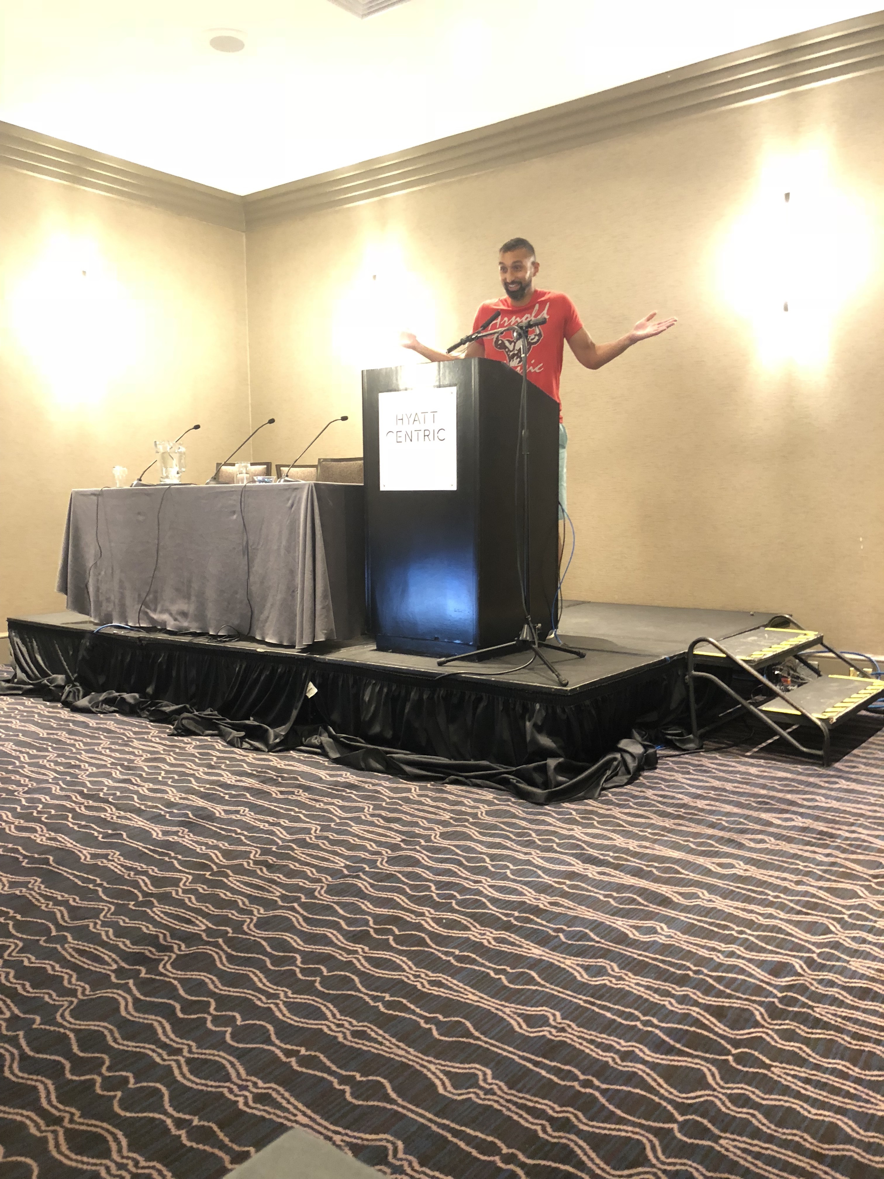 Vivek at a Hyatt Centric podium, arms spread mid-speech, in an empty hotel ballroom