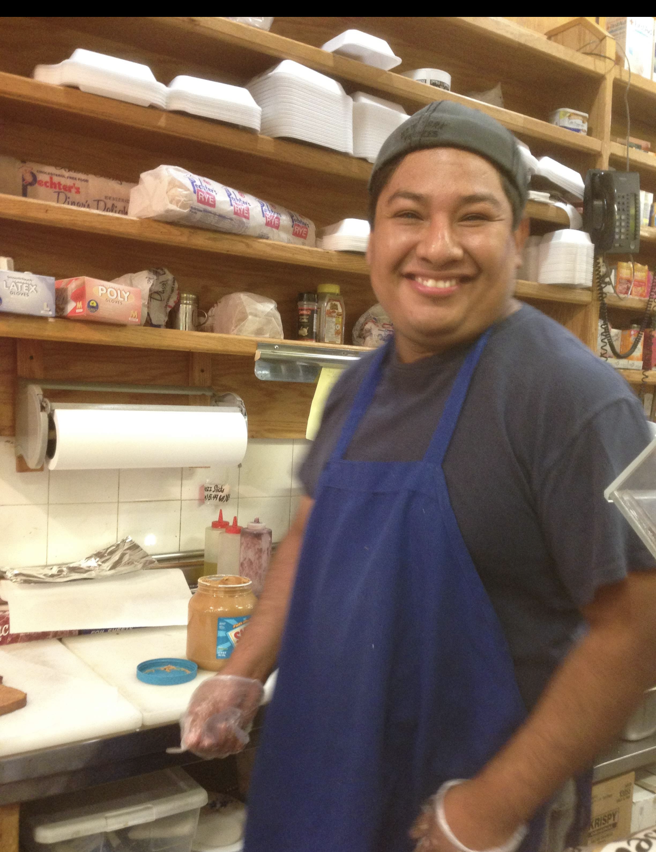 Octavio smiling behind the counter at the Red Pepper deli in a blue apron and gloves, making a sandwich