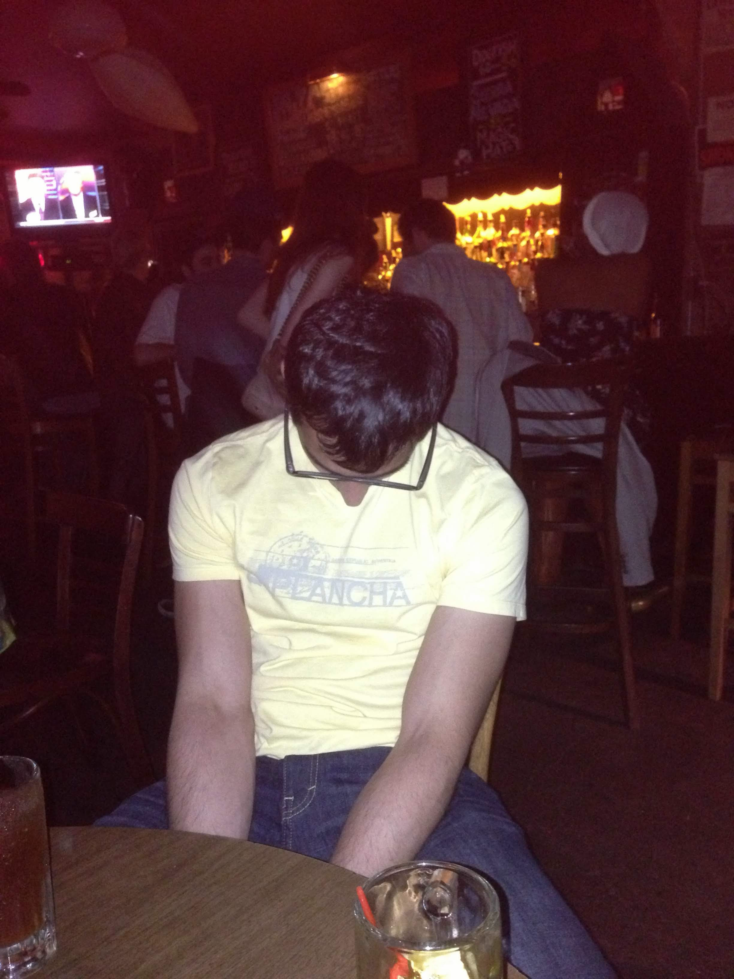 Vivek asleep with his head down at a bar table, drinks in front of him, a busy bar behind