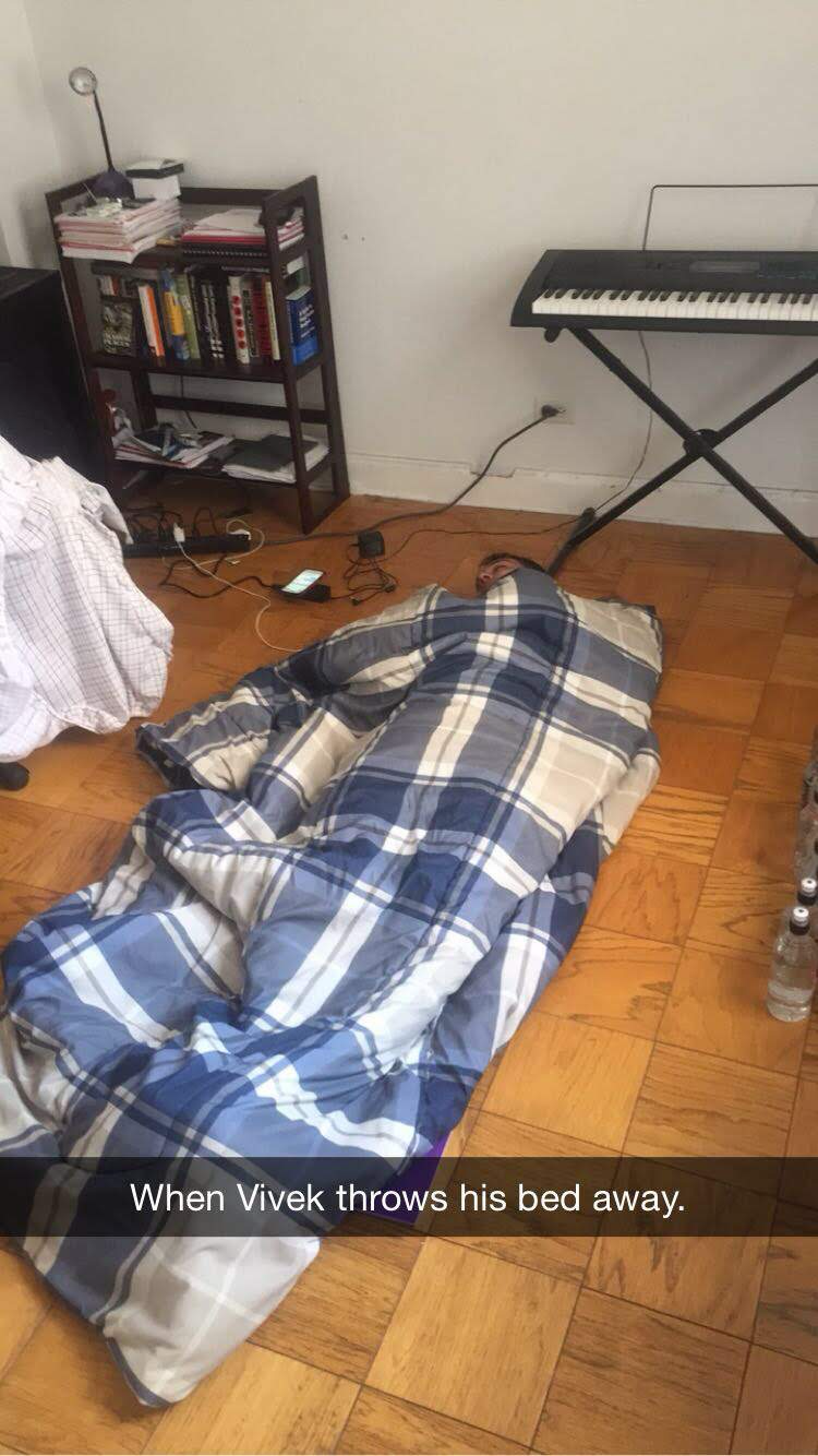Vivek asleep on the hardwood floor of the 7H apartment, wrapped in a plaid comforter with no bed, beside a keyboard and bookshelf